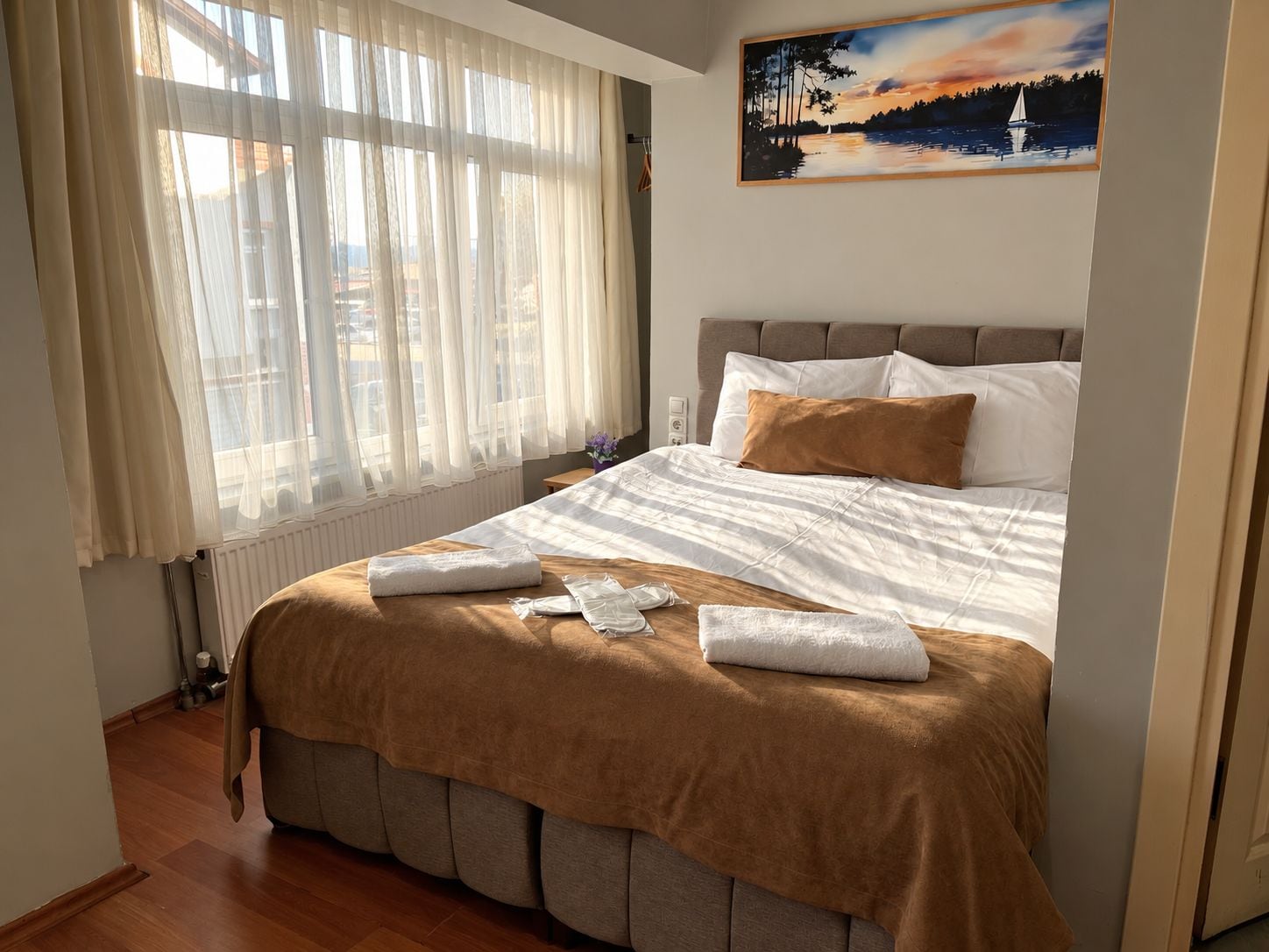 Een hotelkamer met een groot bed, een raam met dunne gordijnen, een schilderij aan de muur en uitzicht op buiten.