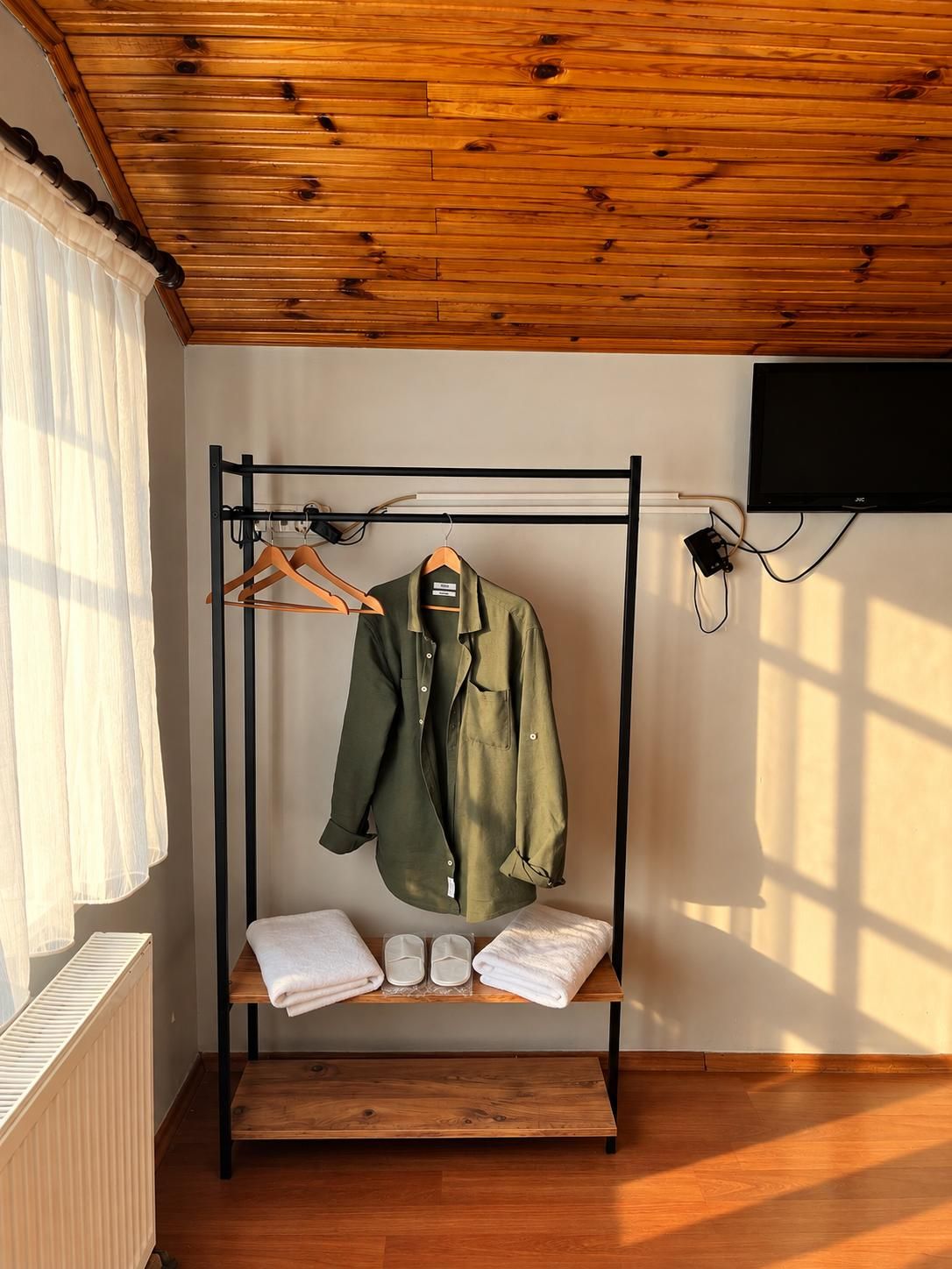 Une pièce avec un plafond en bois, un portant en métal noir où sont suspendus des vêtements et des chaussures, un radiateur blanc et un téléviseur fixé au mur.