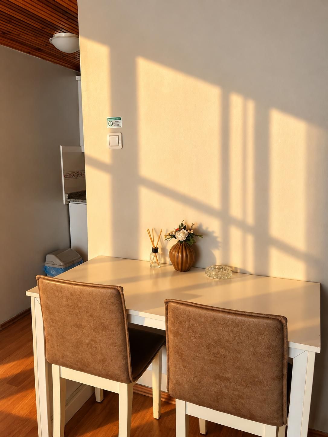 Un comedor con una mesa blanca y dos sillas marrones, un florero con flores, y una mesita con un recipiente de vidrio.