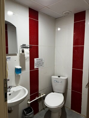 A bathroom with a white sink, a toilet, and a mirror. There is a red and white tiled wall, and a sign with text in a foreign language. - Karaçam Apart Otel (Kastamonu)