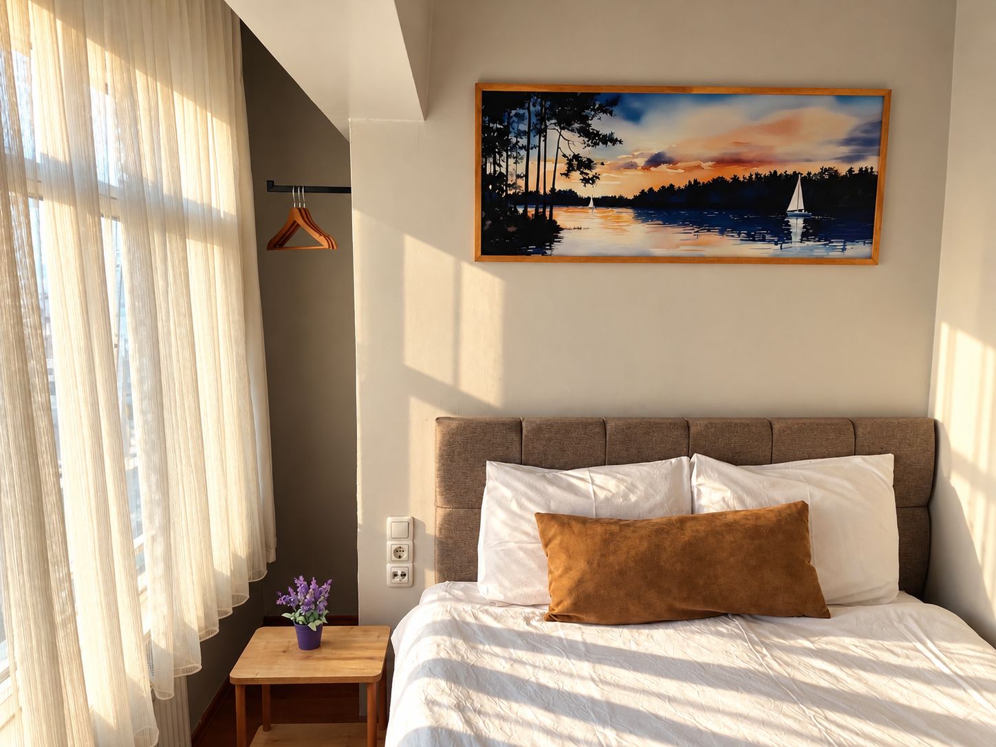 Quarto de hotel com uma cama, pintura de pôr do sol sobre a água, uma mesinha com uma planta e uma janela com cortinas translúcidas.