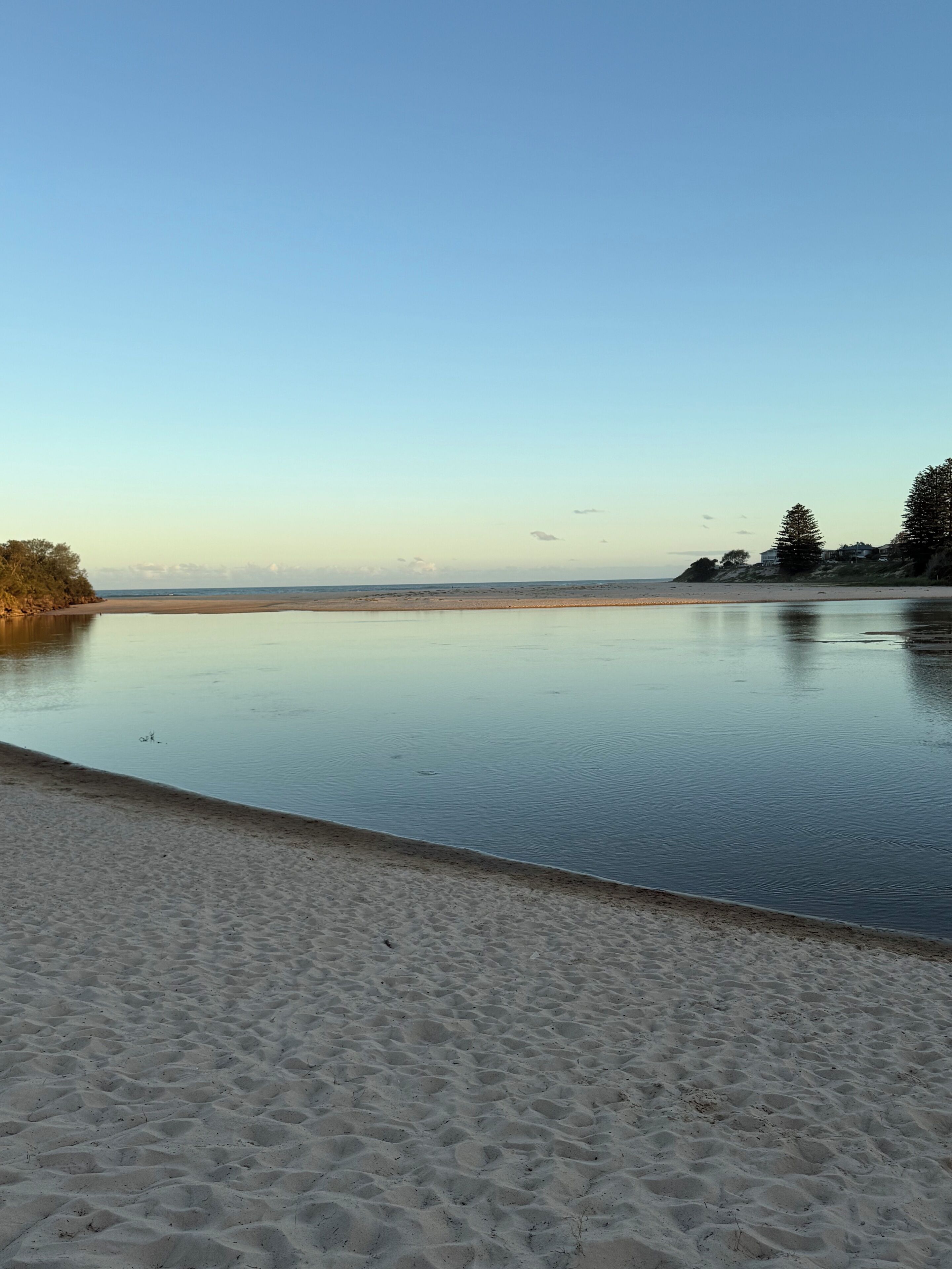 Une plage avec une étendue d’eau calme, un rivage sablonneux et quelques arbres en arrière-plan.