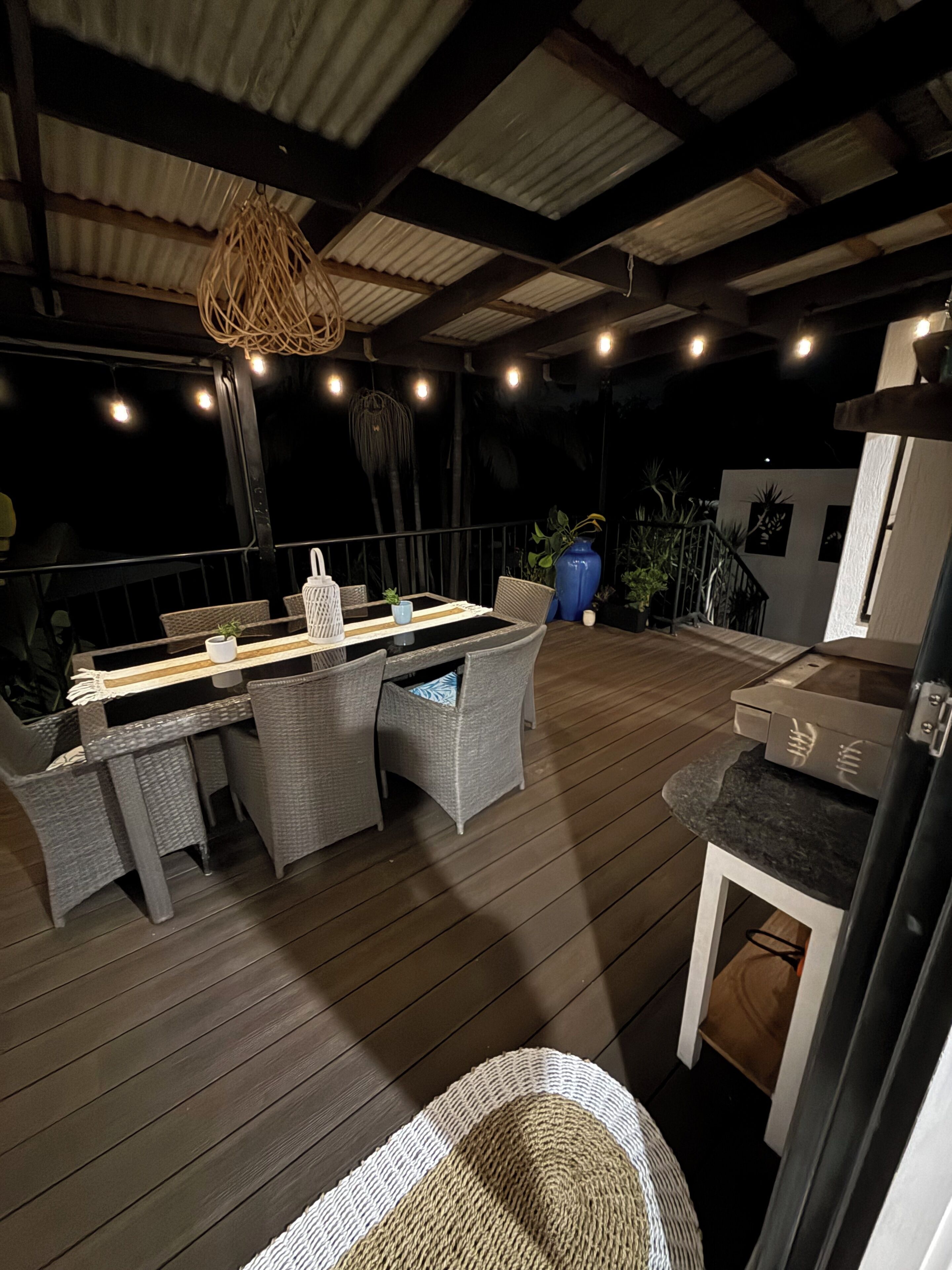 An outdoor patio with a dining table and chairs, surrounded by potted plants and a hanging light fixture.