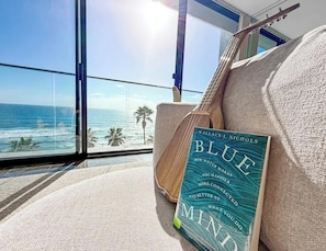 Ein Zimmer mit großem Fenster und Blick auf den Ozean, einer Gitarre und einem Buch mit dem Titel 'Blue Mini'.