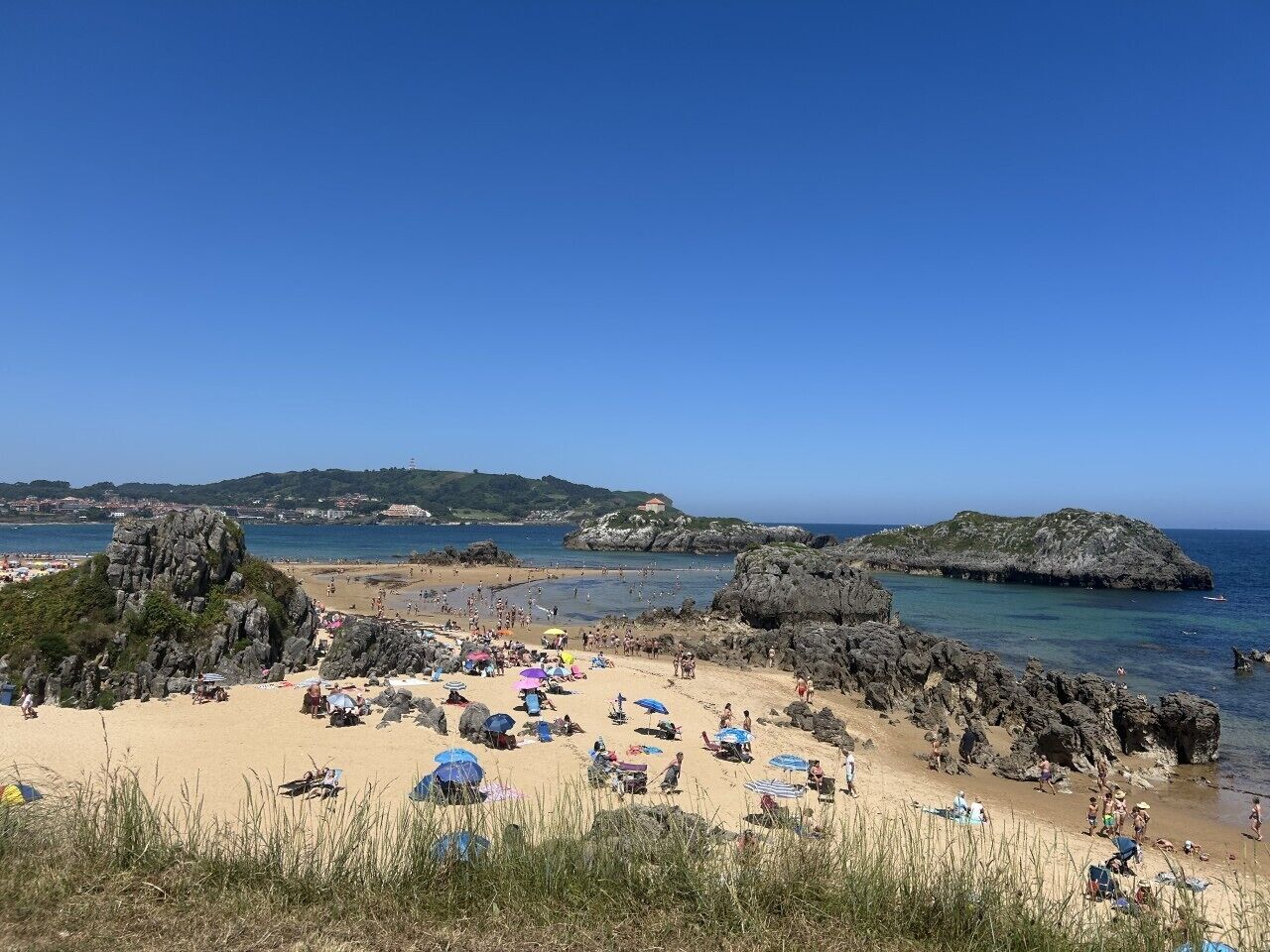 Una playa con formaciones rocosas, cielos despejados y gente disfrutando del sol.
