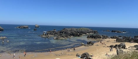 En strand med klippformationer, kristallklart blått vatten och människor som njuter av havet.