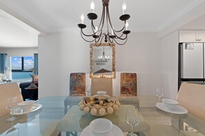 Uma sala de jantar com mesa de vidro, cadeiras, lustre, tigela decorativa e vista para o mar.