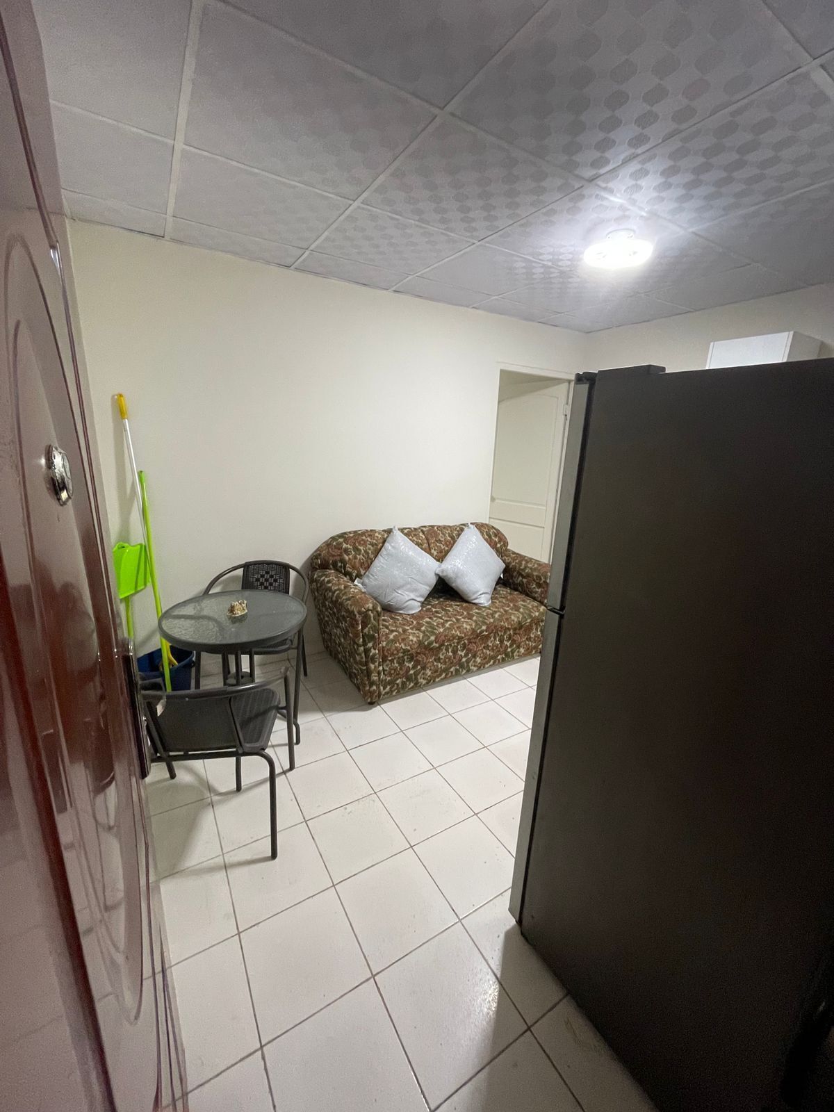 Una habitación compacta con suelo de baldosas, un sofá con cojines, una pequeña mesa redonda con sillas, un refrigerador y una puerta con picaporte.