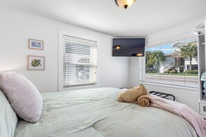 Ein Schlafzimmer mit Bett, zwei Fenstern mit Jalousien, einem wandmontierten Flachbildfernseher und Blick ins Freie.