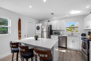 A modern kitchen with white cabinets, stainless steel appliances, a central island with bar stools, and a view of the outdoors through a window. - July Deals - Pet Friendly and Hot tub! Suite B (Indian Rocks Beach)