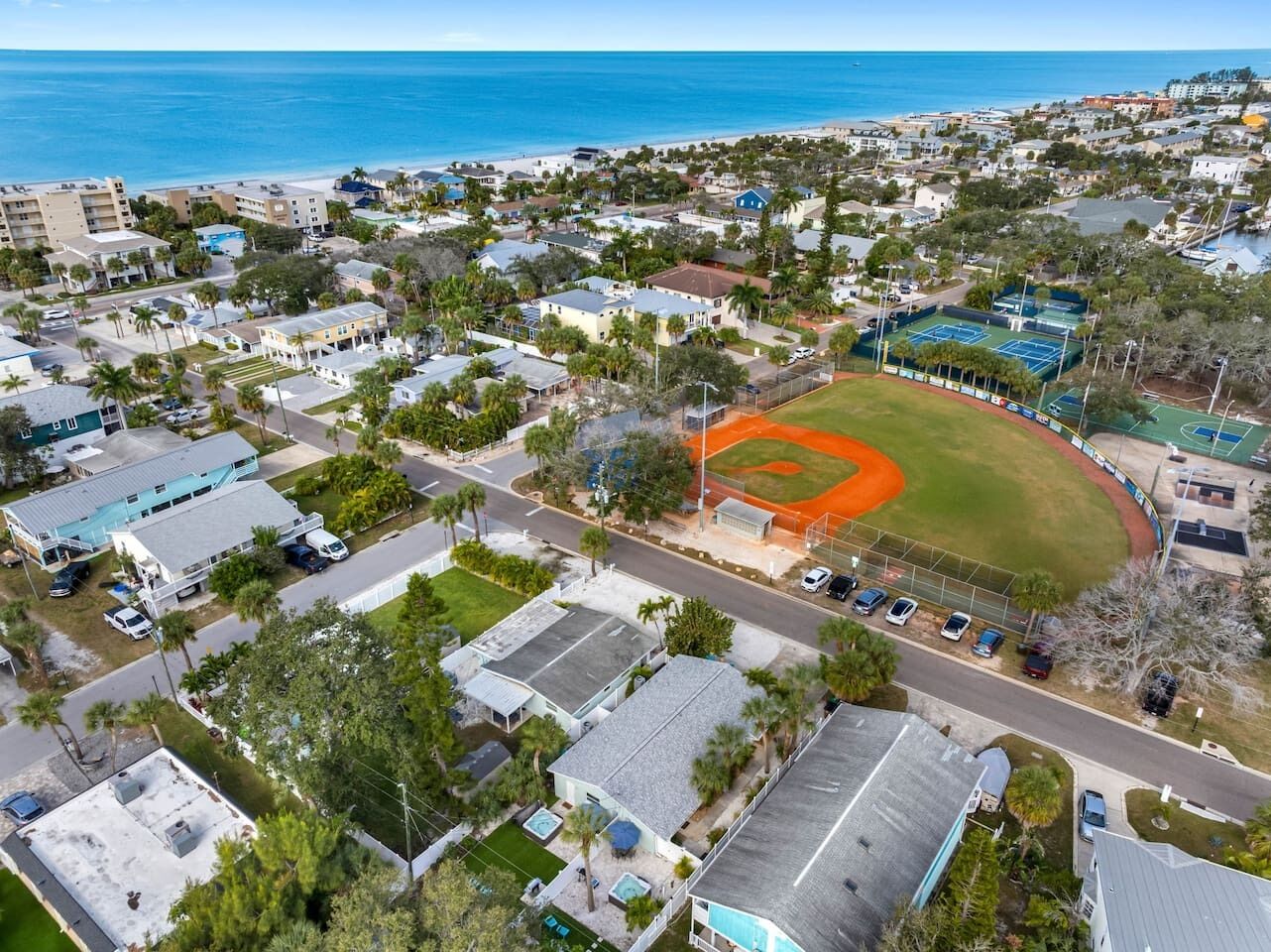 An aerial view of a residential area with houses, a baseball field, and a beach in the background.
