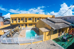 A yellow building with a swimming pool, a fenced patio area, and a playground. - POUSADA VILA ALEGRIA (Penha)