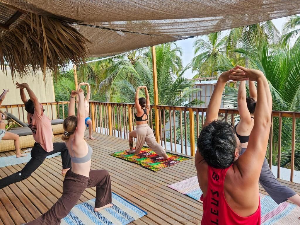 A group of people performing yoga on a wooden deck with a thatched roof, surrounded by palm trees.
