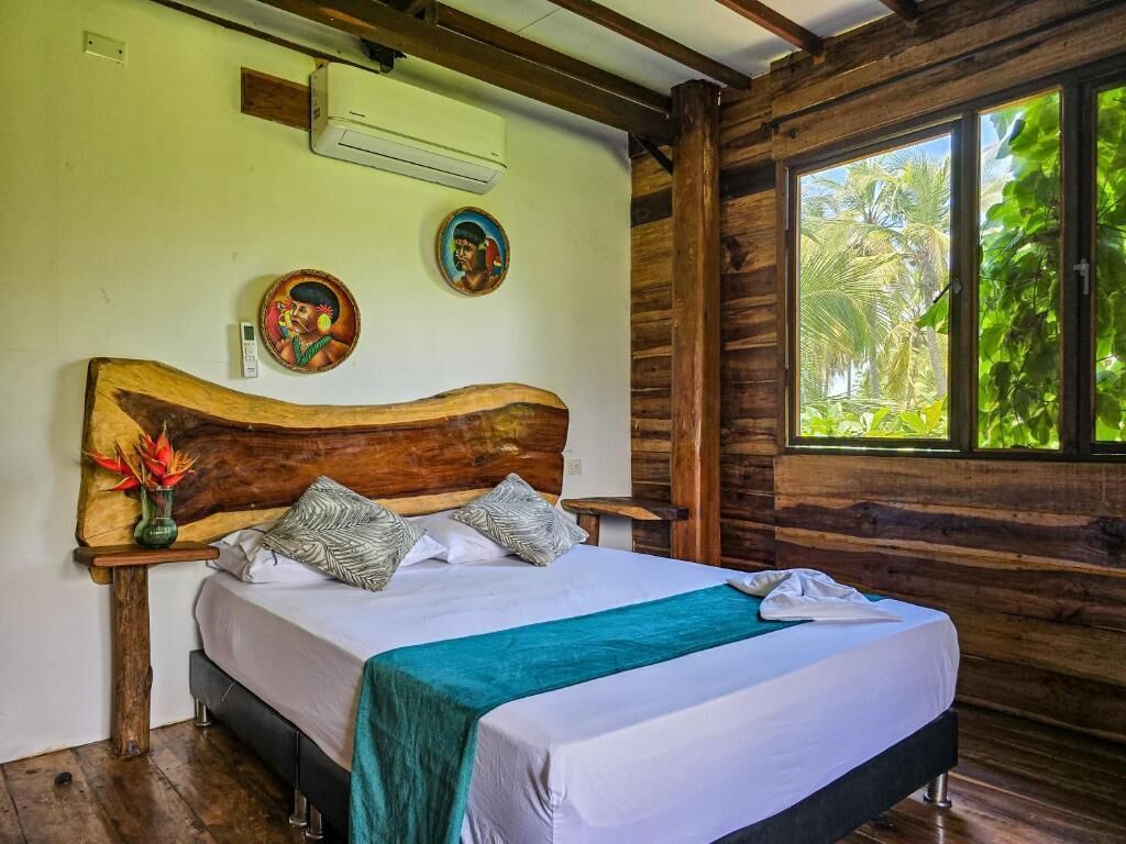 A wooden-framed room with a bed, a wall-mounted air conditioner, and a window with a view of greenery.