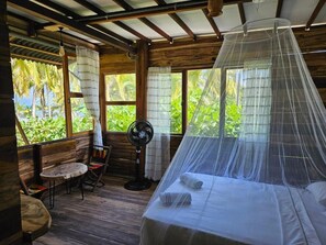 A room with a bed under a mosquito net, a fan, a chair, and large windows with curtains.