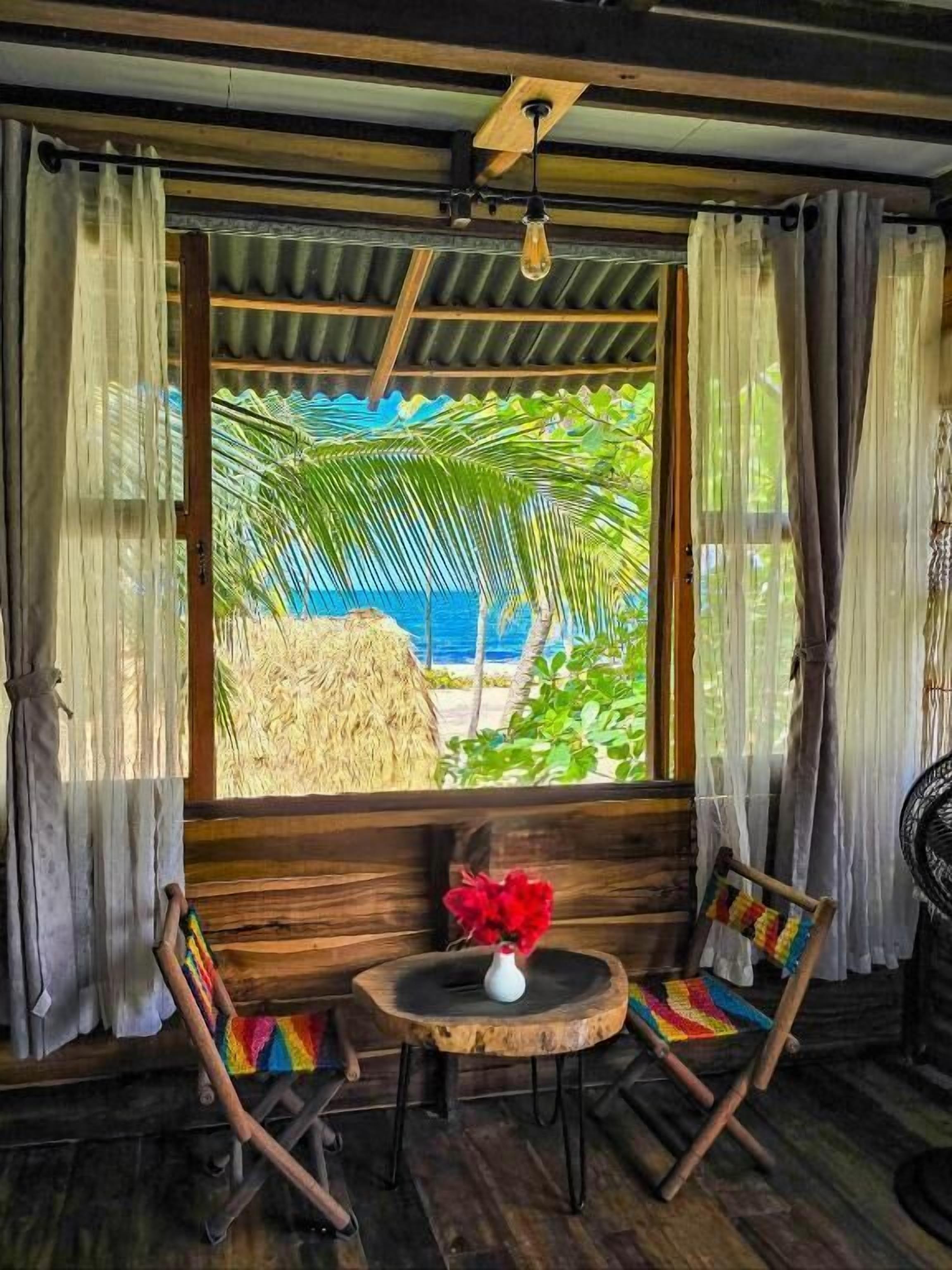 A room with a wooden floor, a round table with a vase of flowers, two wooden chairs, and a view of the beach through a large window with curtains.