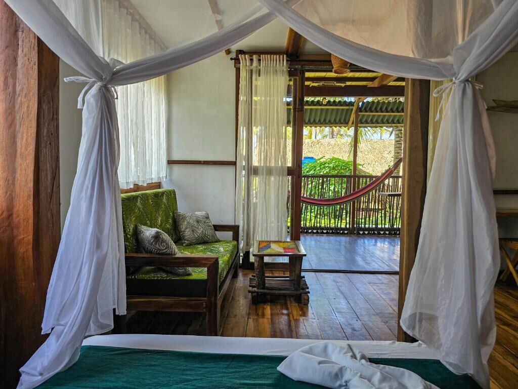 A room with a bed, a small table, and a bench with cushions. There is a view of a wooden deck outside.