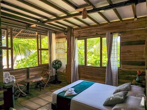 A room with a bed, a fan, a table with towels, and large windows with a view of greenery.