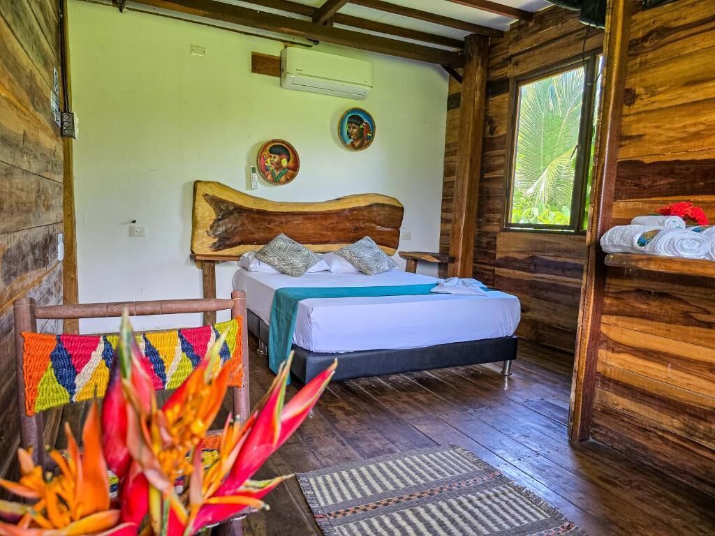 A wooden cabin-style room with a bed, a woven chair, and a window with a view of greenery.