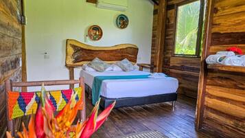 A wooden cabin-style room with a bed, a woven chair, and a window with a view of greenery.