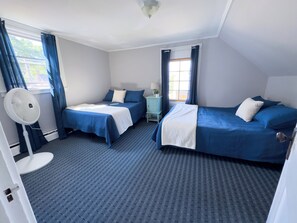 Une chambre d’hôtel avec deux lits, une table de chevet avec une lampe, une fenêtre avec des rideaux et un ventilateur sur pied.