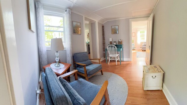 Un salon chaleureux avec un canapé, un fauteuil, une lampe et une petite table. Au fond de la pièce, on aperçoit un coin repas avec une table et des chaises.