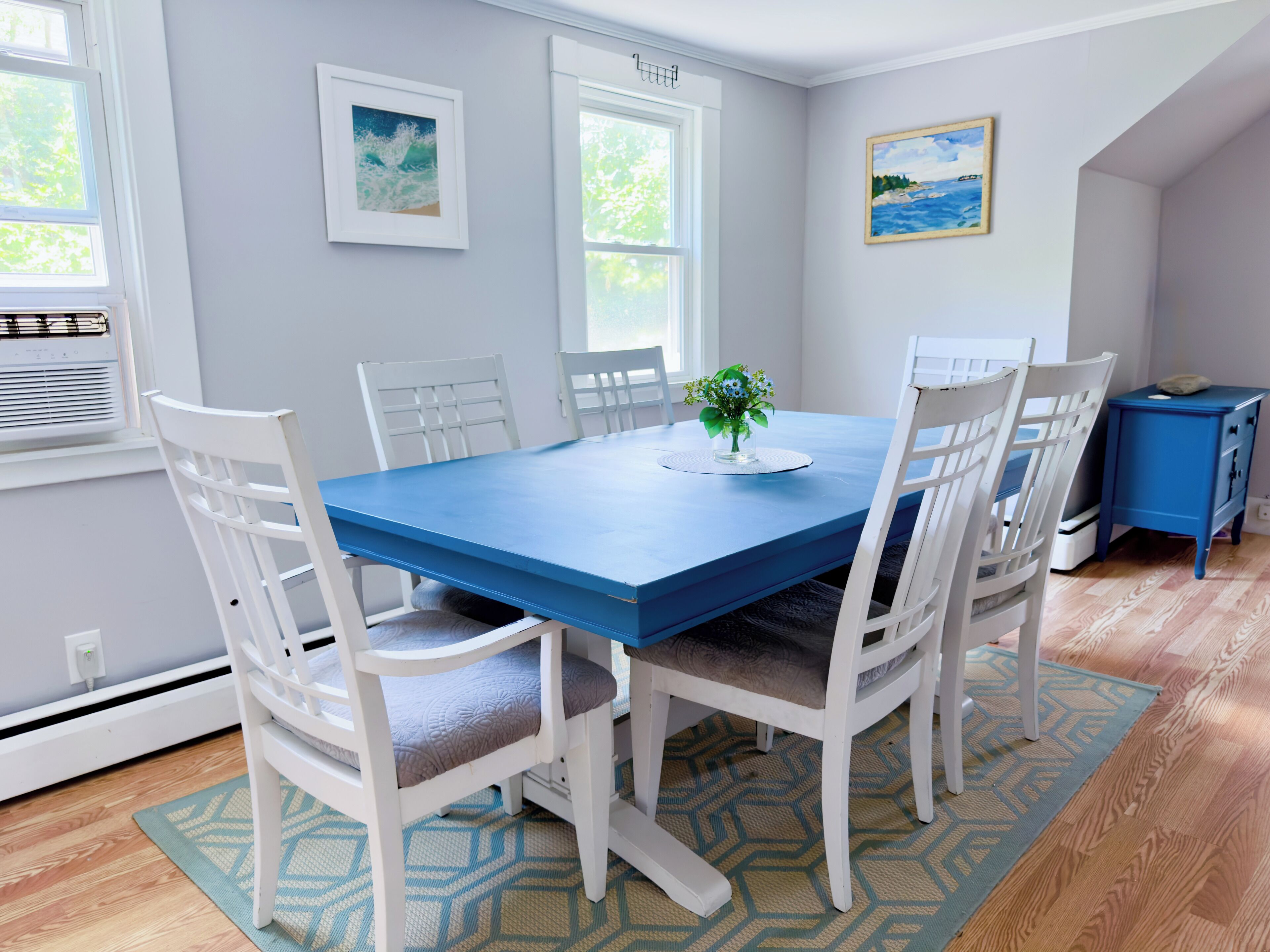 Un coin repas avec une table bleue et des chaises blanches, un petit meuble bleu et deux tableaux encadrés accrochés au mur.