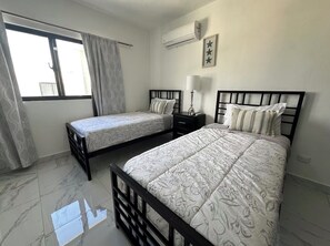 A bedroom with two beds, a window with curtains, a bedside table with a lamp, and a wall-mounted air conditioner. - Spacious 4-bedroom townhome in lovely Punta Cana with AC and free WiFi (Punta Cana)