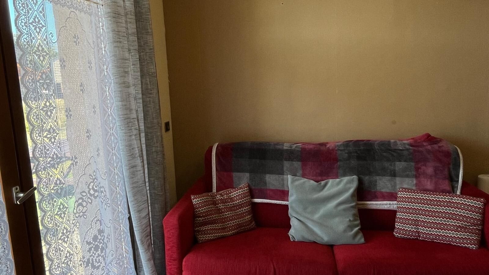 A red sofa with a checkered blanket and patterned pillows, a window with patterned curtains, and a plain wall.