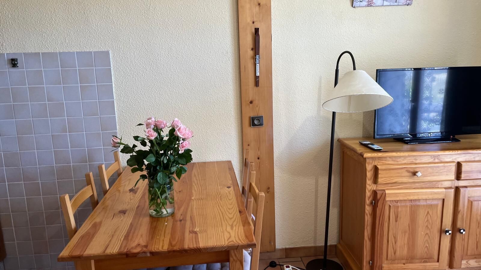 A wooden dining table with a vase of flowers, a television on a wooden cabinet, and a lamp.