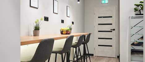 Un área de recepción con un mostrador de madera, cuatro sillas grises, cuadros en la pared, una planta en maceta y una puerta con un cartel.