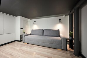 Uma sala de estar moderna com um sofá cinzento, uma pequena mesa lateral com uma planta e um candeeiro fixado na parede.