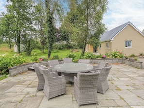 A patio with a round table and wicker chairs, surrounded by a stone wall and greenery.