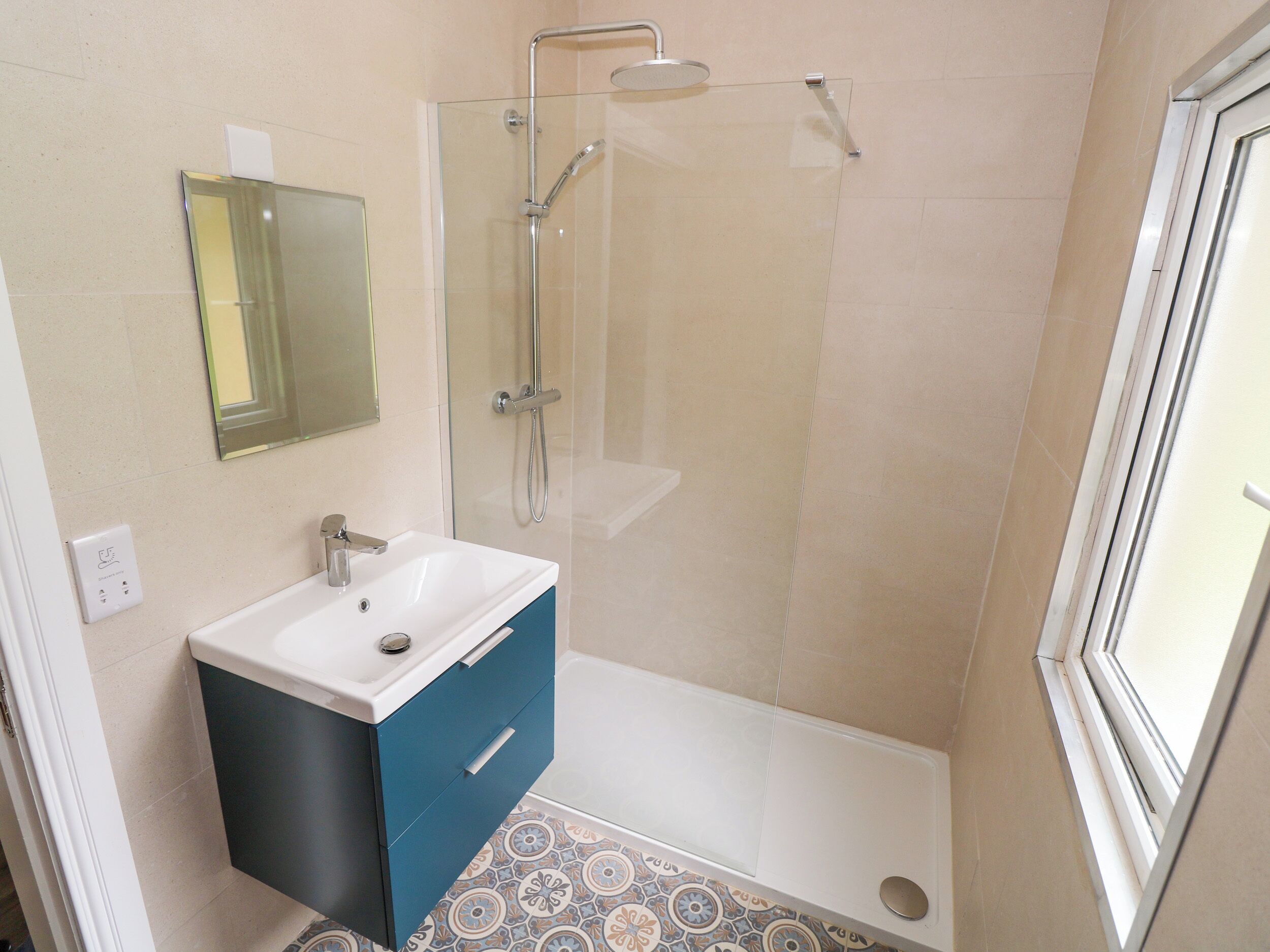 A bathroom with a glass shower enclosure, a white sink with a blue cabinet, and patterned floor tiles.