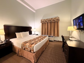 A hotel room with a large bed, a flat-screen TV, a desk with a lamp, and a window with curtains.