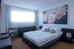 A bedroom with a bed, a chair, a nightstand, a lamp, and a large painting on the wall. - Lux Modern 2BR Flat wth Balcony • Walk to Battersea Park, River, Mall, Transport (London)