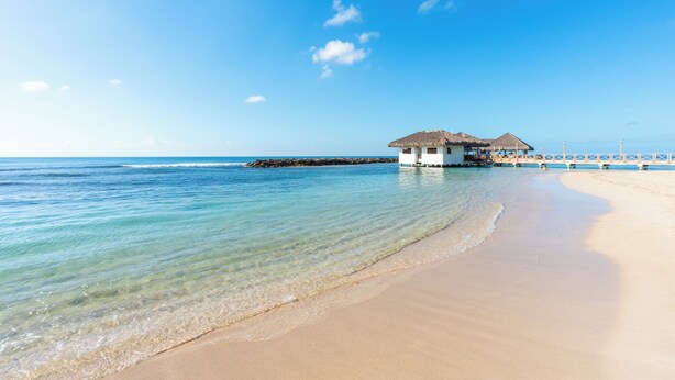 Una playa con agua cristalina, un edificio de estilo bungalow y un muelle que se adentra en el mar.
