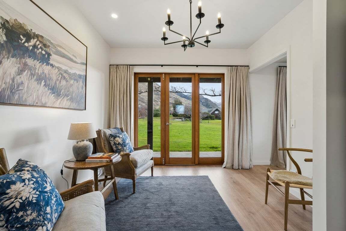 A modern living room with a sofa, a side table, a lamp, and a large painting. There is a view of a green landscape through a glass door.