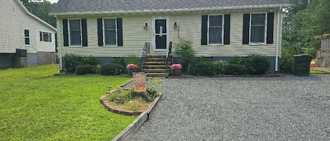 A two-story house with a front porch, a gravel driveway, and a small garden with pink flowers.