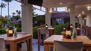 Een terras met houten tafels en stoelen, lantaarns en uitzicht op palmbomen en gebouwen.