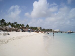 A beach with clear water, palm trees, and a few buildings in the background. - Rare OCEANFRONT- Beautiful 2 Bed-Marriott Aruba Surf Club - Full Resort Access (Noord)