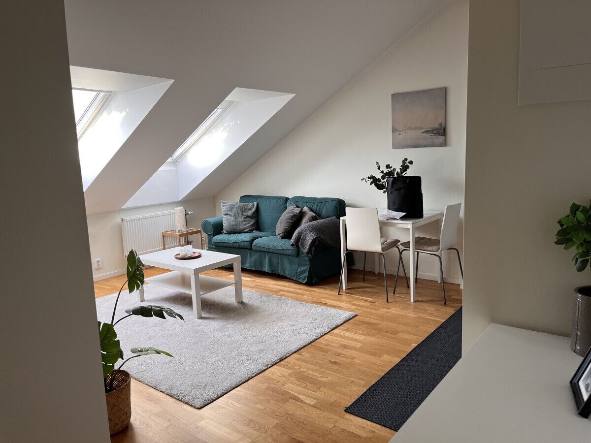A modern living room with a sofa, a coffee table, and a dining table. There are skylights on the ceiling, a plant, and a painting on the wall.
