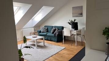 A modern living room with a sofa, a coffee table, and a dining table. There are skylights on the ceiling, a plant, and a painting on the wall.