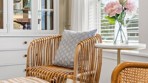 A wicker chair with a patterned cushion, a small round table with a vase of flowers, and a window with blinds. - South Haven Retreat • Indoor/Outdoor Pool • Beach (South Haven)