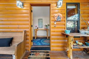 A wooden cabin interior with a wooden bench, a small table, a rug, and a door leading to a room with a bed and a wall-mounted sign. - Trade the Heat for the Pines: Pinetop Family Cabin, Kayaks & Cool Nights (Pinetop)