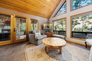 A cozy interior with a stone fireplace, a gray sofa, a wooden coffee table, and large windows offering views of the outdoors. - Trade the Heat for the Pines: Pinetop Family Cabin, Kayaks & Cool Nights (Pinetop)