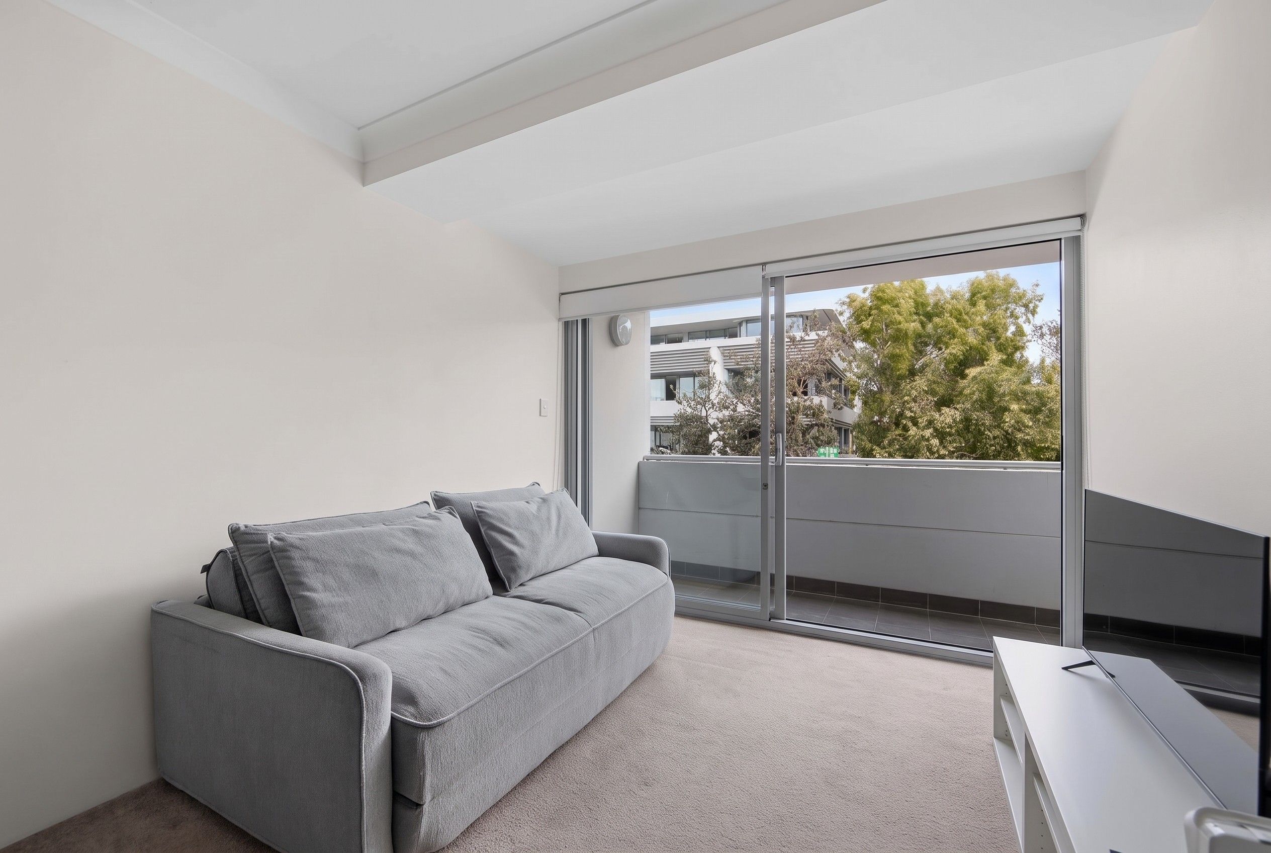 En moderne lejlighed med en grå sofa, et lille skrivebord og en stor skydedør, der fører ud til en balkon med udsigt over træer og bygninger.