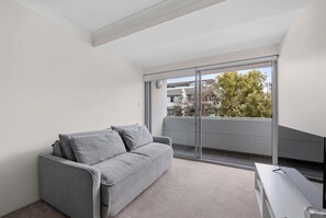 Ein modernes Apartment mit einem grauen Sofa, einem kleinen Schreibtisch und einer großen Schiebeglasdoor zum Balkon mit Blick auf Bäume und Gebäude.