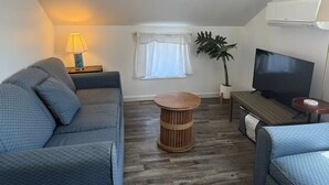 A living room with a sofa, a TV, a lamp, a plant, and a round wooden table. - Ocean Glass | Family Cottage | Pet-Friendly Stay | Steps to Sand (Rehoboth Beach)