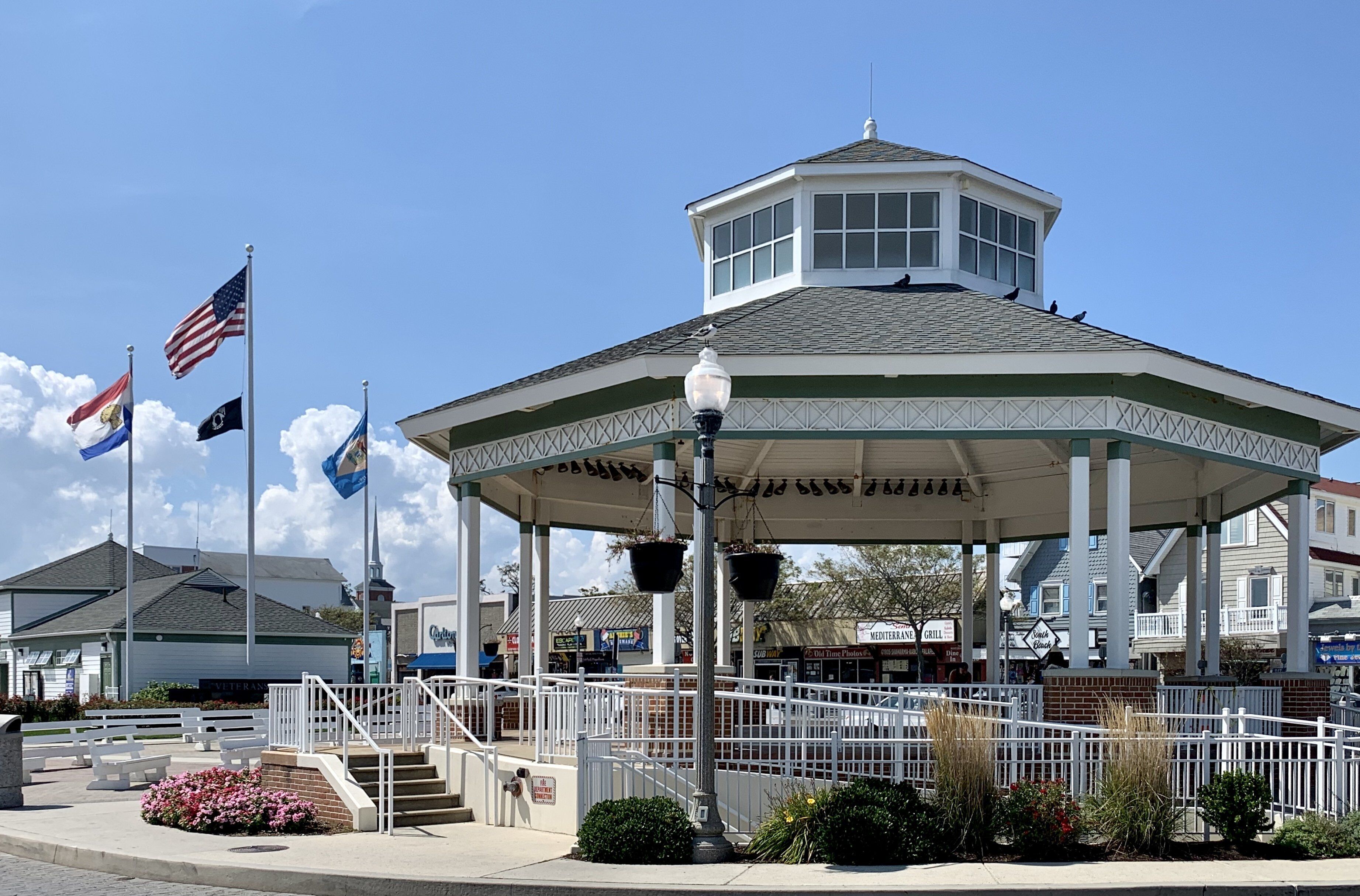 A gazebo with a central dome and a surrounding railing, located in a public space with flags and benches.